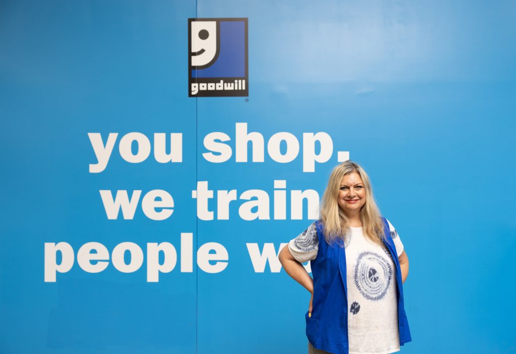 Debbie stands in front of a blue wall at a Goodwill store that reads "you shop, we train, people work."